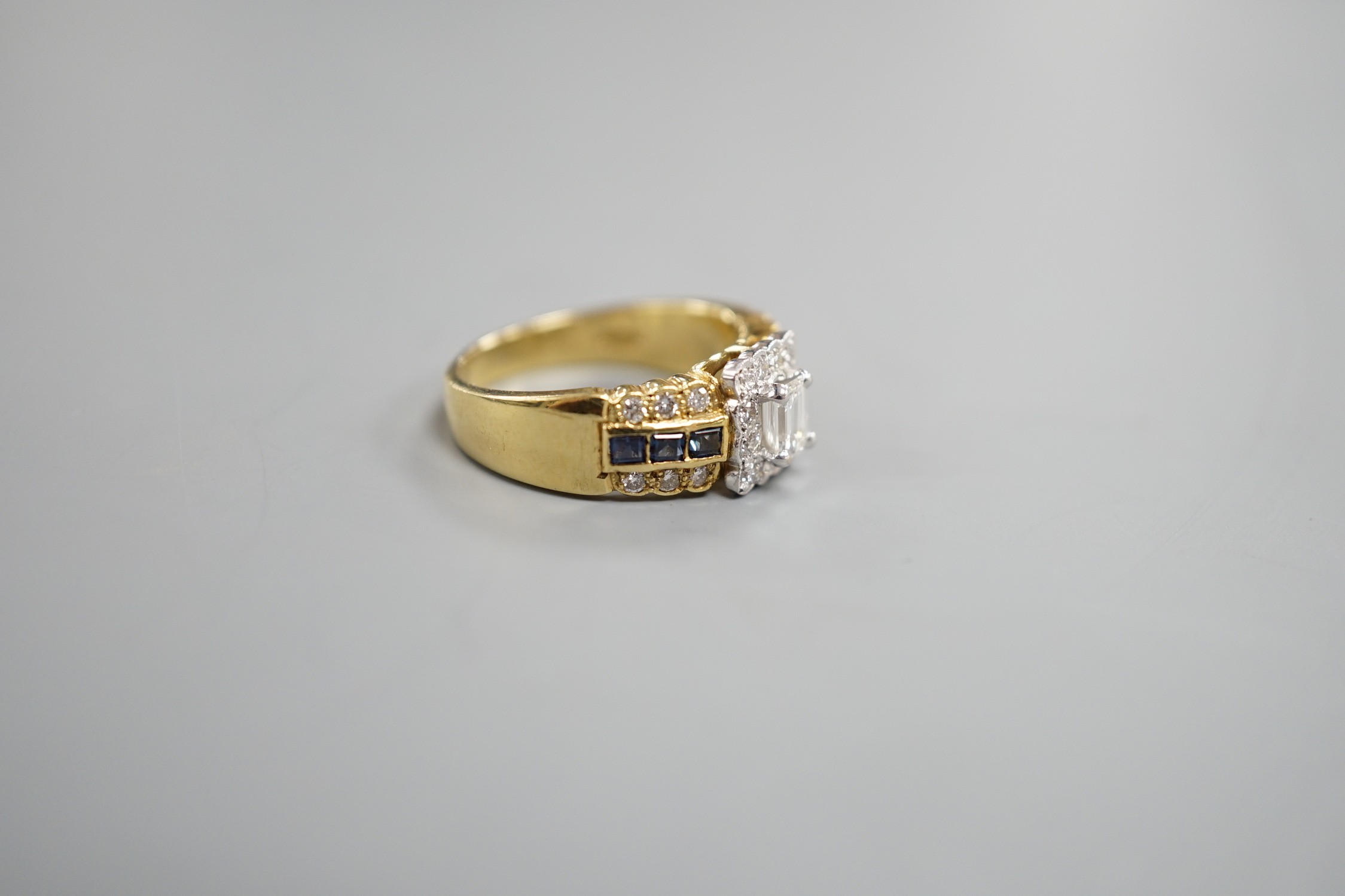 A modern 18ct gold, emerald and round cut diamond set square cluster ring, three three row sapphire and diamond set shoulders, size M, gross weight 6.6 grams.